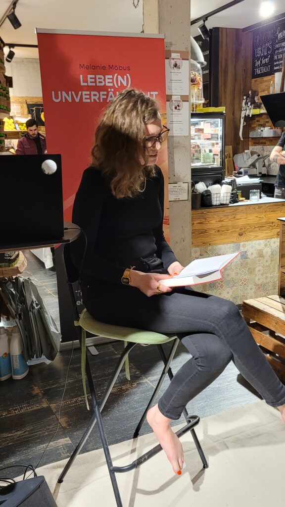 Lebe(n) unverfälscht im Bioladen: Melanie Möbus liest barfuß auf einem Stuhl sitzend aus ihrem Buch vor.